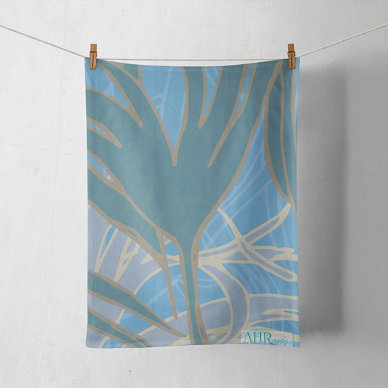 Colourful gift – Blue, Turquoise and Sand Kelp seaweed design tea towel hanging from clothesline, shadows showing on off-white background. Designed by Anne Harrington Rees. Designed, printed and made in Ireland.