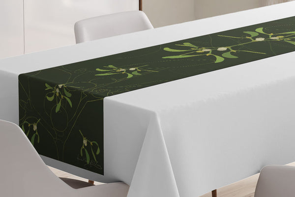 Close up of the Mistletoe table runner designed by Anne Harrington Rees, viewed on a table covered with a white tablecloth.  Symmetrical design of leaves and berries on a dark green background.