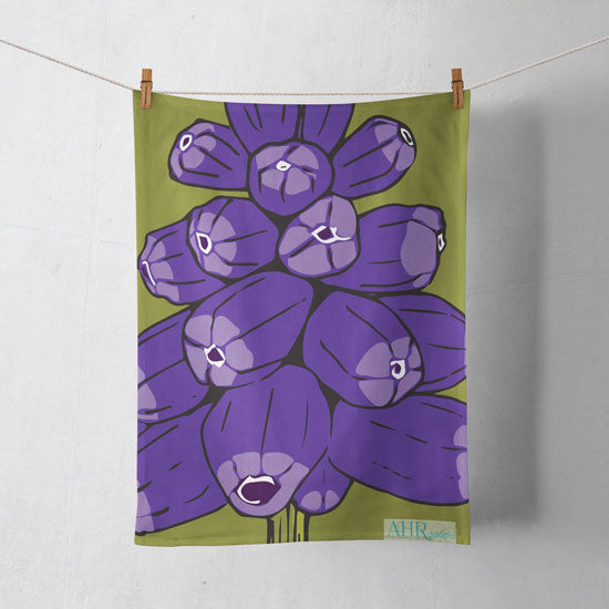 Colourful gift – Purple and Green Muscari flower design tea towel hanging from clothesline, shadows showing on off-white background. Designed by Anne Harrington Rees. Designed, printed and made in Ireland.