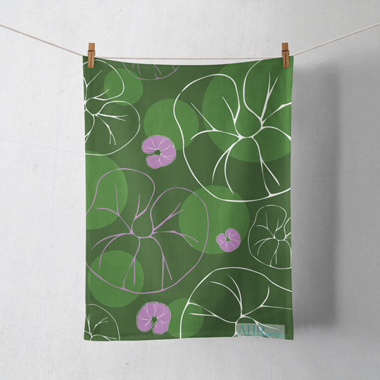 Colourful gift – Green, Pink and White Sea Bindweed flower design tea towel hanging from clothesline, shadows showing on off-white background. Designed by Anne Harrington Rees. Designed, printed and made in Ireland.
